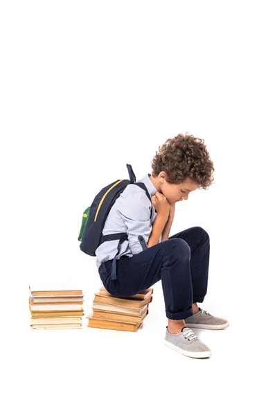 Sconvolto scolaro con zaino seduto su libri isolati su bianco — Foto stock