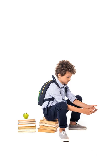 Curly schoolboy with backpack sitting on books near apple and using smartphone isolated on white — Stock Photo