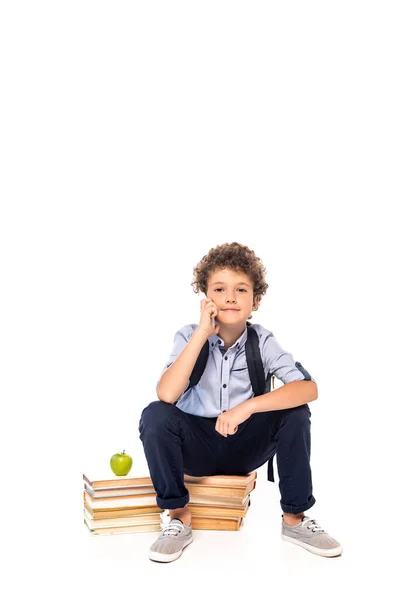 Lockiger Schuljunge mit Rucksack sitzt auf Büchern in der Nähe von Apfel und spricht auf Smartphone isoliert auf weiß — Stockfoto