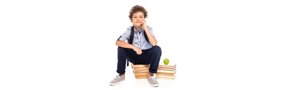 Panoramic shot of schoolboy with backpack sitting on books near apple and talking on smartphone isolated on white — Stock Photo