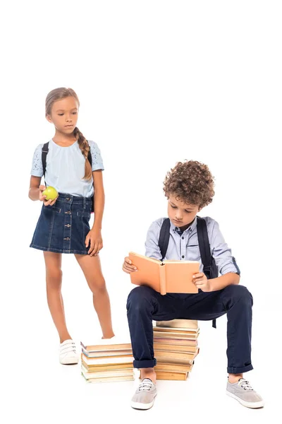 Школьница держит яблоко и смотрит на мальчика, сидящего на книгах и читающего изолированные на белом — стоковое фото