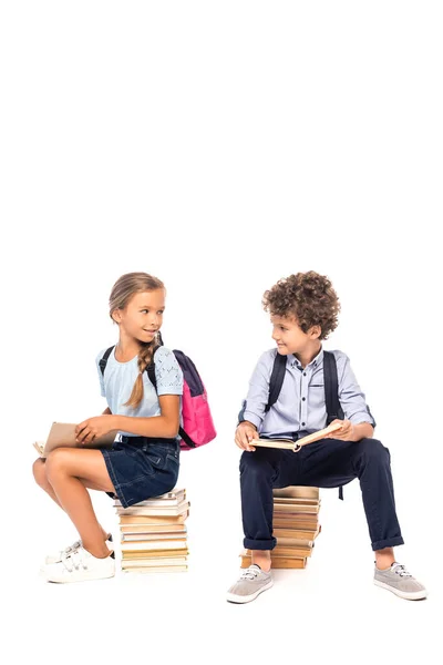 Schoolkids sitting on books and looking at each other isolated on white — Stock Photo