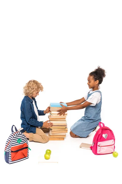 Мультикультурні діти сидять і зворушливі книги біля стиглих яблук і рюкзаків ізольовані на білому — стокове фото