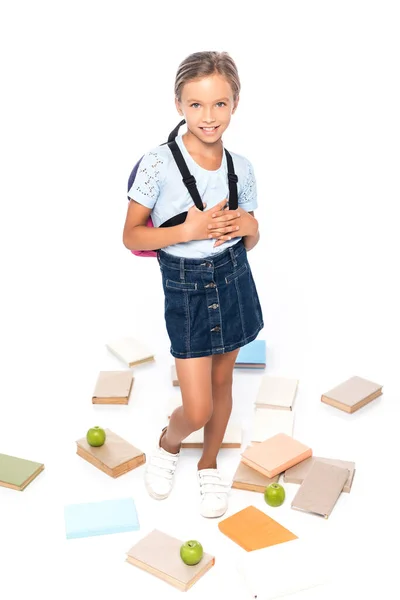 Colegiala con mochila de pie cerca de manzanas y libros aislados en blanco - foto de stock