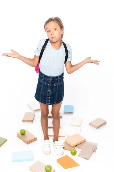 Colegiala con mochila de pie cerca de manzanas y libros, mientras que muestra gesto de encogimiento aislado en blanco - foto de stock