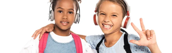 Cultivo panorámico de escolares escuchando música en auriculares inalámbricos, abrazando a un amigo afroamericano y mostrando tres dedos aislados en blanco - foto de stock