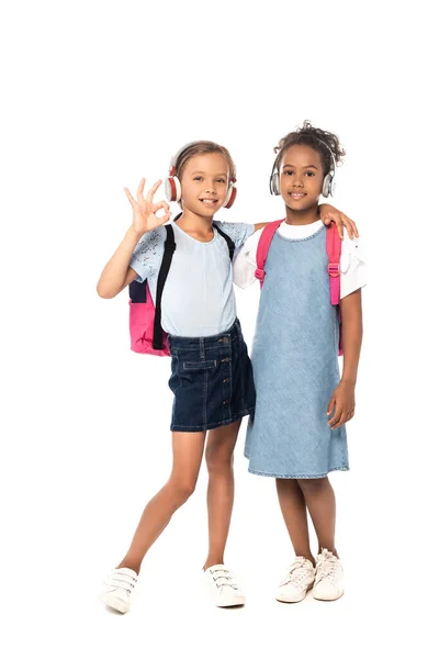 Schoolkid listening music in wireless headphones, hugging african american friend and showing ok sign isolated on white — Stock Photo