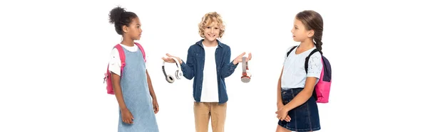 Panoramic shot of blonde schoolboy holding wireless headphones near multicultural schoolgirls isolated on white — Stock Photo