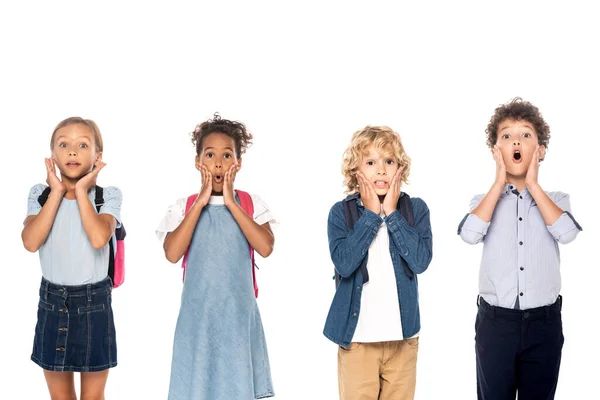 Shocked multicultural schoolgirls and schoolboys touching faces while looking at camera isolated on white — Stock Photo