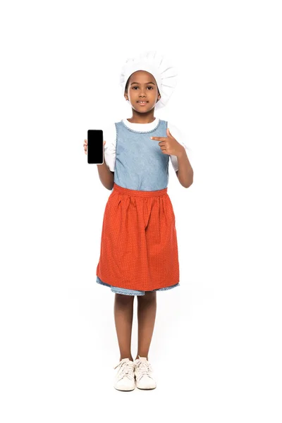 Afrikanisch-amerikanisches Kind im Kostüm des Kochs zeigt mit dem Finger auf Smartphone mit leerem Bildschirm isoliert auf weiß — Stockfoto