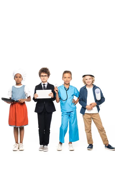 Niños multiculturales en trajes de diferentes profesiones que sostienen sartén, plano, estetoscopio y tableta digital aislados en blanco - foto de stock
