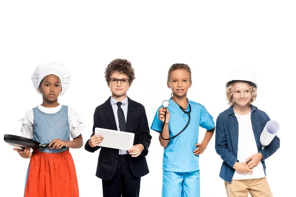 Enfants multiculturels en costumes de différentes professions tenant plan, poêle à frire, stéthoscope et tablette numérique isolés sur blanc — Photo de stock