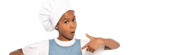Panoramic crop of surprised african american kid in costume of chef pointing with finger isolated on white — Stock Photo