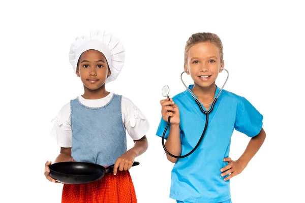 Multicultural kids in costumes of different professions holding frying pan and stethoscope isolated on white — Stock Photo