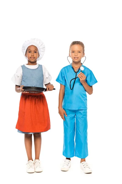 Мультикультурные дети в костюмах разных профессий держат сковородку и стетоскоп стоя изолированными на белом — стоковое фото