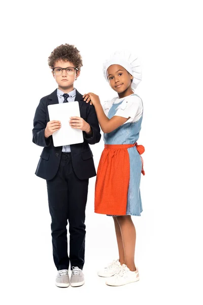 Niño afroamericano tocando chico en gafas y traje sosteniendo tableta digital aislado en blanco - foto de stock