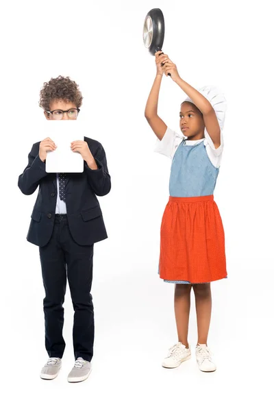 Afro-américain enfant prétendant femme au foyer et tenant poêle près du garçon dans des lunettes couvrant le visage avec tablette numérique isolé sur blanc — Photo de stock