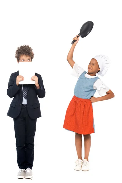 African american kid pretending housewife and holding frying pan near boy in suit covering face with digital tablet isolated on white — Stock Photo