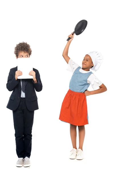 Niño afroamericano fingiendo ama de casa y sosteniendo sartén cerca de niño en traje cubriendo la cara con tableta digital aislada en blanco - foto de stock