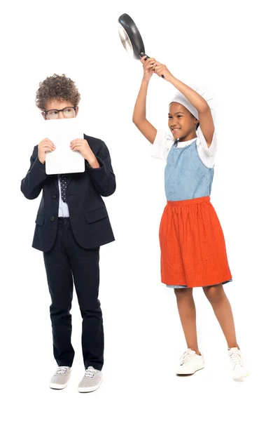 Afro-américaine enfant prétendant femme au foyer et tenant poêle près du garçon en costume couvrant le visage avec tablette numérique isolé sur blanc — Photo de stock