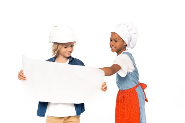 Niño rizado en casco de seguridad y traje de arquitecto mirando el plano cerca de niño afroamericano en sombrero de chef aislado en blanco - foto de stock