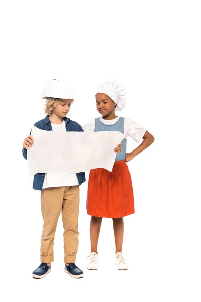 Кудрявый мальчик в защитном шлеме и костюме архитектора, смотрящего на чертеж рядом с африканским американским ребенком в шляпе шеф-повара, стоящим с рукой на бедре, изолированным на белом — стоковое фото