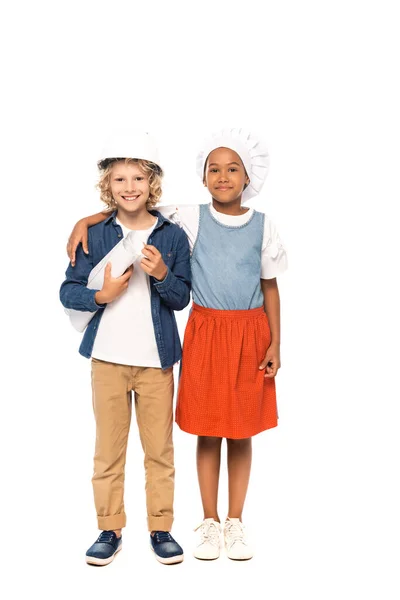 Afro-américain enfant en chapeau de chef étreignant garçon bouclé dans un casque de sécurité et costume d'architecte debout avec un plan isolé sur blanc — Photo de stock