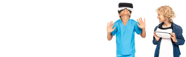 Панорамная концепция кудрявого мальчика, смотрящего на ребенка в наушниках виртуальной реальности, жестикулирующего изолированно на белом — стоковое фото