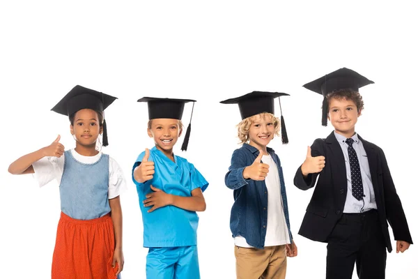 Crianças multiculturais em bonés de graduação vestidos com trajes de diferentes profissões mostrando polegares isolados em branco — Fotografia de Stock