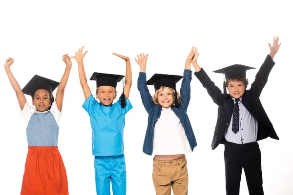 Niños multiculturales en gorras de graduación vestidos con disfraces de diferentes profesiones de pie con las manos levantadas aisladas en blanco - foto de stock