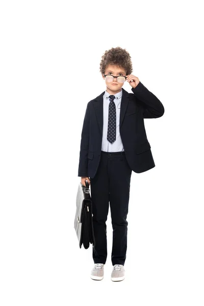 Curly boy in suit touching glasses and holding briefcase isolated on white — Stock Photo