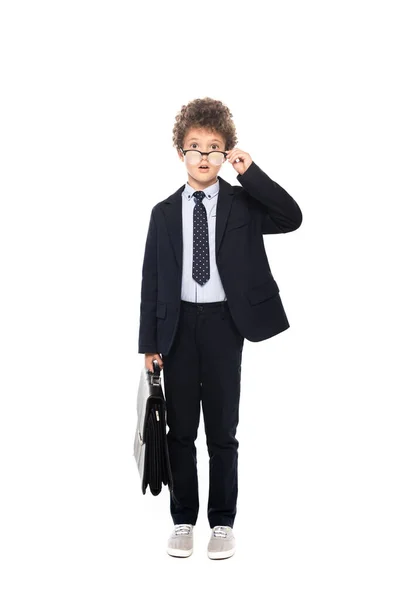 Niño sorprendido en traje tocando gafas y sosteniendo maletín aislado en blanco - foto de stock