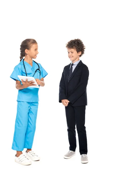 Ребенок в костюме врача, держащего цифровой планшет возле кудрявого мальчика в формальной одежде изолированы на белом — стоковое фото