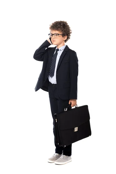 Niño rizado en traje y gafas que sostiene el maletín mientras habla en el teléfono inteligente aislado en blanco - foto de stock