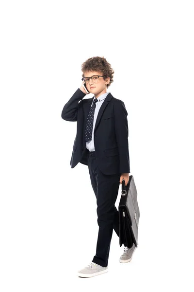 Niño con traje y gafas sosteniendo el maletín mientras habla en el teléfono inteligente aislado en blanco - foto de stock