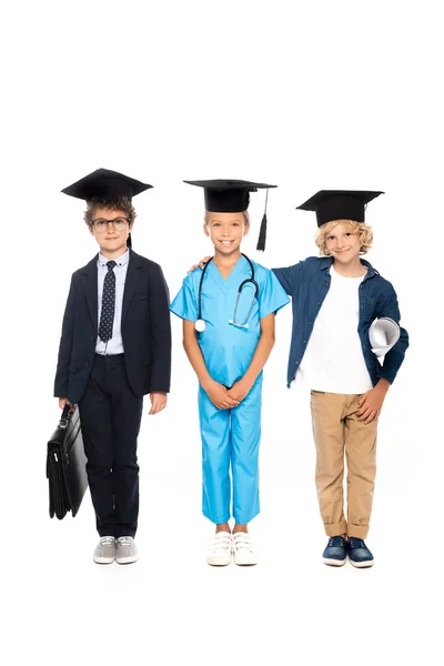 Enfants en casquettes de graduation vêtus de costumes de différentes professions debout avec stéthoscope, plan et mallette isolés sur blanc — Photo de stock