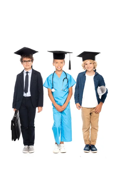 Niños en gorras de graduación vestidos con disfraces de diferentes profesiones de pie con estetoscopio, anteproyecto y maletín aislado en blanco - foto de stock