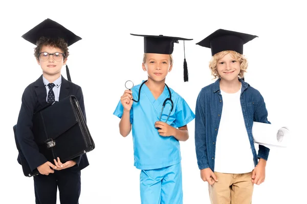 Children in graduation caps dressed in costumes of different professions holding blueprint, stethoscope and briefcase isolated on white — Stock Photo