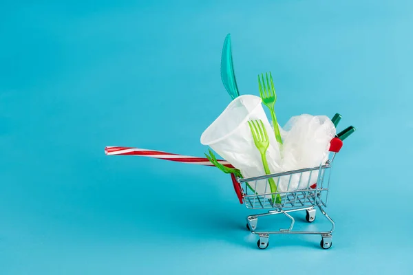 Objetos de plástico desechables en un pequeño carrito de compras sobre fondo azul - foto de stock