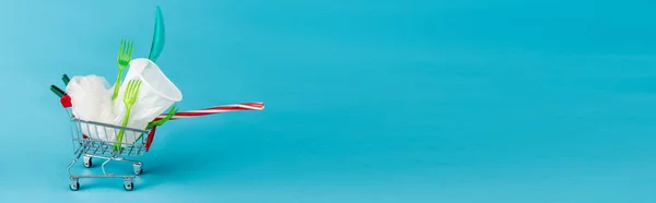 Disposable plastic objects in small shopping cart on blue background, panoramic shot — Stock Photo