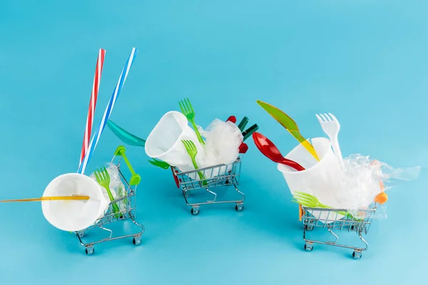 Objetos de plástico desechables en pequeños carritos de compras sobre fondo azul - foto de stock
