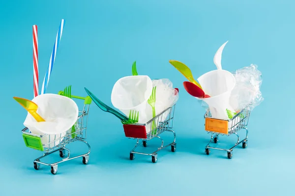 Objets jetables en plastique dans de petits chariots sur fond bleu — Photo de stock