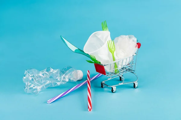 Objetos de plástico desechables en un pequeño carrito de compras sobre fondo azul - foto de stock