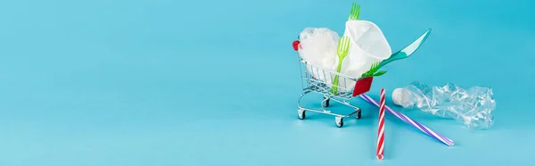 Objetos de plástico desechables en un pequeño carrito de compras sobre fondo azul, plano panorámico - foto de stock