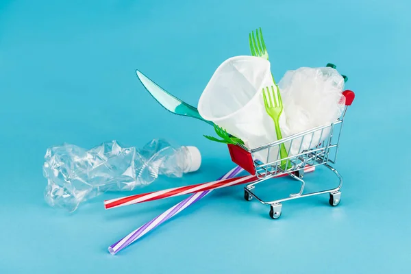 Objetos de plástico desechables en un pequeño carrito de compras sobre fondo azul - foto de stock