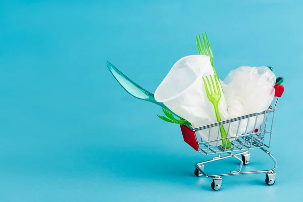 Objetos de plástico desechables en un pequeño carrito de compras sobre fondo azul - foto de stock