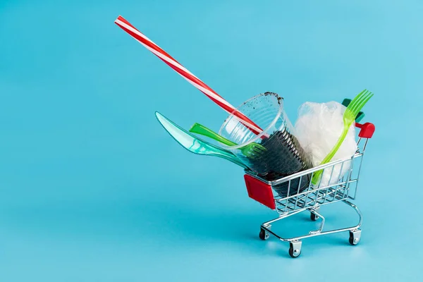 Objetos de plástico desechables y molidos en un pequeño carrito de compras sobre fondo azul - foto de stock