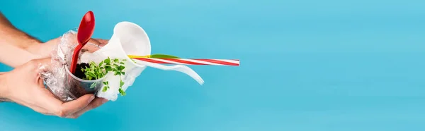Cropped view of man holding plastic objects and green seedling on blue background, panoramic shot — Stock Photo