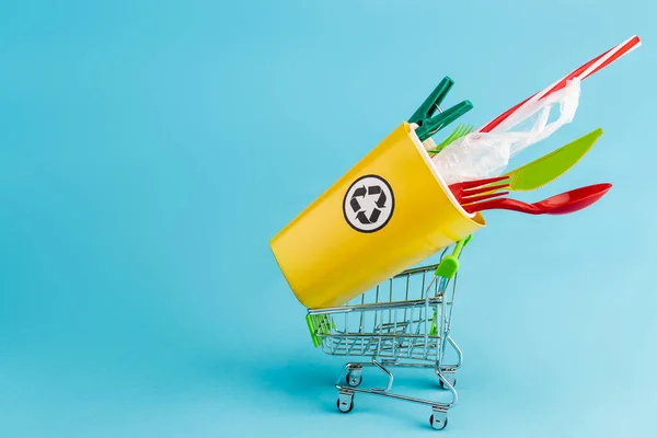 Papelera de reciclaje amarillo con plástico en un pequeño carrito de compras sobre fondo azul - foto de stock