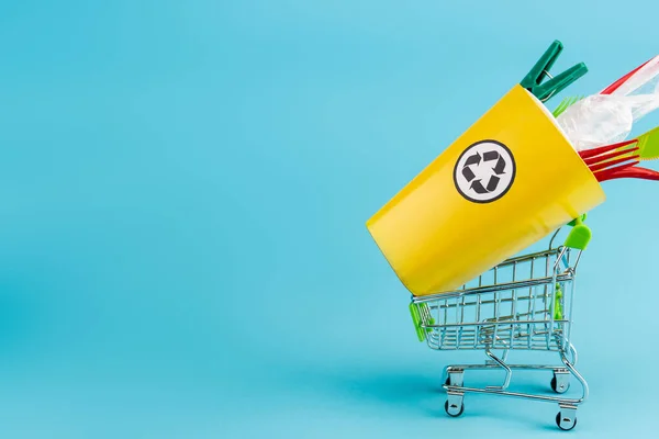 Papelera de reciclaje amarillo con plástico en un pequeño carrito de compras sobre fondo azul - foto de stock